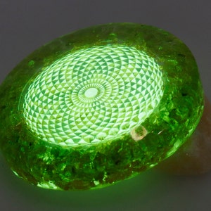 May include: A glowing green, oval-shaped decorative object. The center features a detailed, geometric pattern that emits a bright light. The outer edge has a textured, rough appearance. The object is resting on a light-colored surface.
