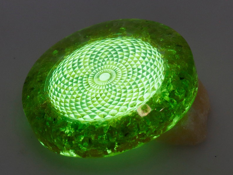 May include: A glowing green, oval-shaped decorative object. The center features a detailed, geometric pattern that emits a bright light. The outer edge has a textured, rough appearance. The object is resting on a light-colored surface.