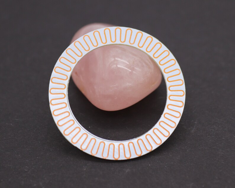 May include: A white circular bezel with a copper-colored ring of small ovals around the edge.