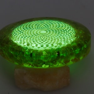 May include: A glowing green decorative object with a geometric pattern on top. The object is round with a textured edge and sits on a light-colored, irregular base. The top surface has a detailed, illuminated design.