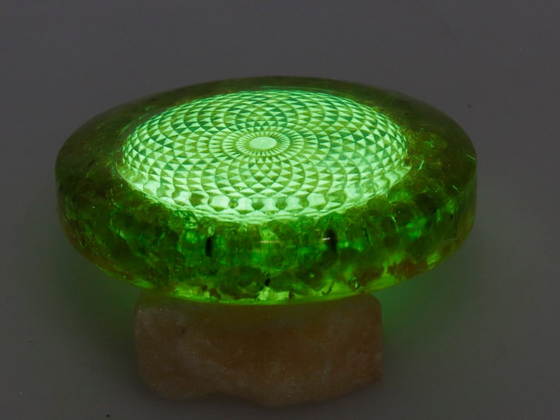 May include: A glowing green decorative object with a geometric pattern on top. The object is round with a textured edge and sits on a light-colored, irregular base. The top surface has a detailed, illuminated design.