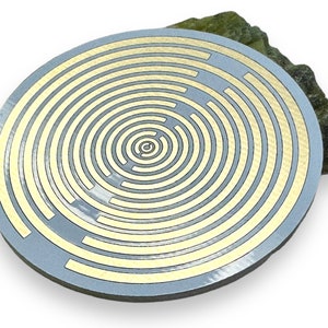 Puede incluir: Un componente electrónico circular de color plateado con un patrón concéntrico de trazas conductoras doradas. El componente está diseñado para la carga inalámbrica.