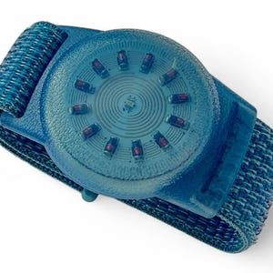 Peut inclure: Bracelet bleu avec un cadran circulaire texturé. Le cadran comporte une série de petites lumières rouges sur le pourtour. Le bracelet est en tissu bleu tissé. L'appareil semble être électronique.