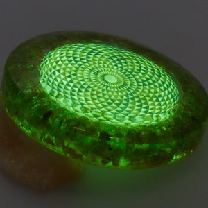 May include: A glowing green, oval-shaped object with a geometric design in the center. The object has a textured, translucent appearance, with a lighter green, patterned interior. The object is resting on a beige, irregular-shaped base.