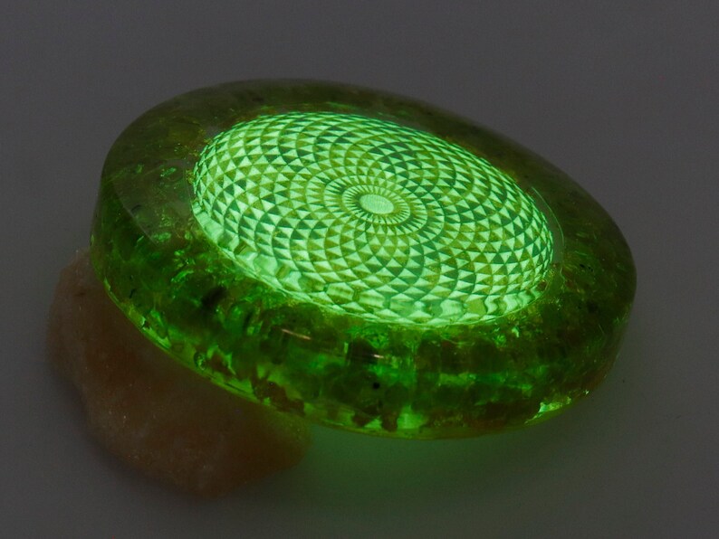 May include: A glowing green, oval-shaped object with a geometric design in the center. The object has a textured, translucent appearance, with a lighter green, patterned interior. The object is resting on a beige, irregular-shaped base.