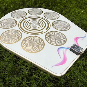 May include: White, semi-circular device with gold spiral patterns and a central circular design. Features a black connector and a pink and blue abstract design. The device is set against a backdrop of green foliage.