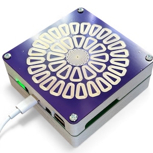 May include: A square electronic device featuring a purple top panel with a gold-colored circular pattern of repeating geometric shapes and a central spiral. The unit has a light-colored side casing, four corner screws, and a white USB cable plugged in.