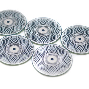 May include: Five round, silver-colored coasters with a blue and white geometric pattern. The coasters have a circular design in the center.