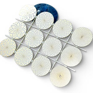Può includere: Un gioco decorativo di tris con pedine circolari argentate e bianche con un motivo floreale. Le pedine sono disposte su una griglia con linee argentate. Sullo sfondo c'è una pietra blu a forma ovale.