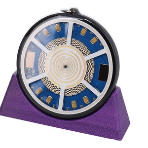Può includere: Un dispositivo elettronico circolare con un design a circuito stampato blu e oro, esposto su un supporto triangolare viola. Il dispositivo presenta un motivo a spirale ed è racchiuso in una cornice nera.