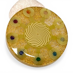 May include: A round, golden-colored orgone energy device with embedded gemstones. The device features a central geometric pattern and radiating spirals, with various colored stones set around the perimeter. The base has a wooden appearance.