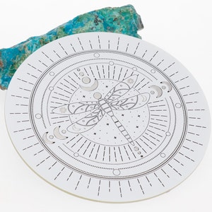 Può includere: Un sottobicchiere circolare bianco con un design in bianco e nero di una libellula circondata da un cerchio di lune e stelle. Il sottobicchiere ha un design a raggiera attorno al bordo.