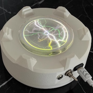 Può includere: Un espositore circolare in plastica bianca con un disco centrale in vetro che si illumina con motivi al plasma elettrico verdi e bianchi. La base include un interruttore a levetta in metallo, una porta di alimentazione e un cavo USB-C bianco.
