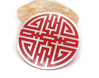 May include: A round, silver-colored metal charm with a red, intricate design. The design is a traditional Chinese symbol known as the "double happiness" symbol.