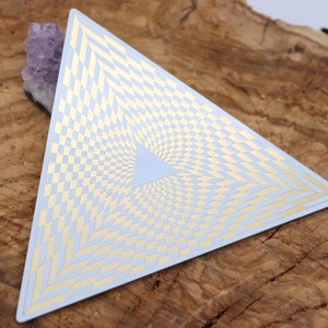 Puede incluir: Una tarjeta triangular blanca con un patrón geométrico dorado que crea una ilusión óptica de un vórtice giratorio.