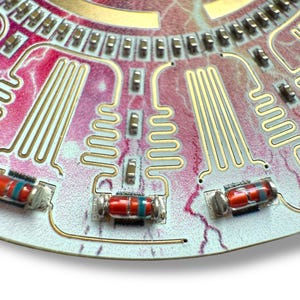 May include: Close-up of a circuit board with gold-colored wiring and red and blue resistors. The board has a pink and white background.