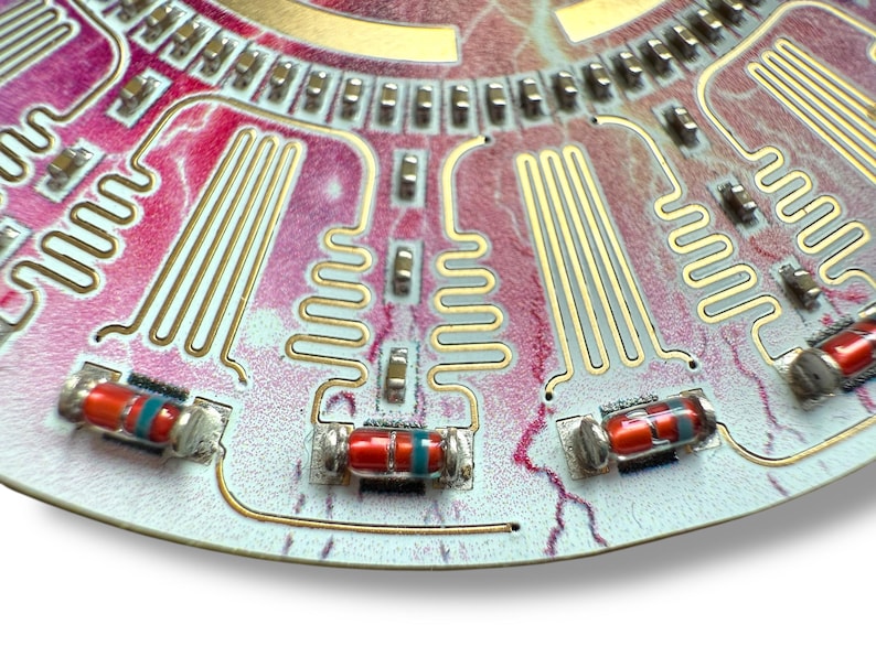 May include: Close-up of a circuit board with gold-colored wiring and red and blue resistors. The board has a pink and white background.