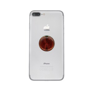 May include: A silver smartphone with a dual-lens camera system and an Apple logo on the back. A circular, copper-coloured accessory with a concentric ring pattern is adhered to the centre of the phone's rear casing, positioned above the text iPhone.