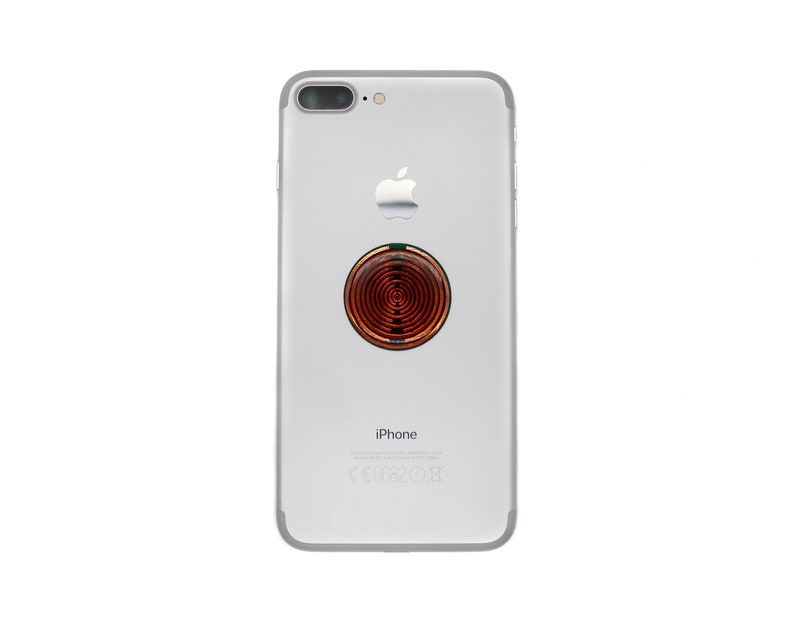 May include: A silver smartphone with a dual-lens camera system and an Apple logo on the back. A circular, copper-coloured accessory with a concentric ring pattern is adhered to the centre of the phone's rear casing, positioned above the text iPhone.