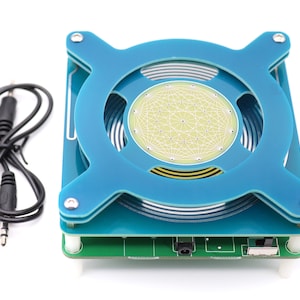 Könnte beinhalten: Eine blaue und grüne Platine mit einer kreisförmigen Spule und einem kleinen weißen Kreis mit einem geometrischen Muster in der Mitte. Die Platine hat ein schwarzes Audiokabel.