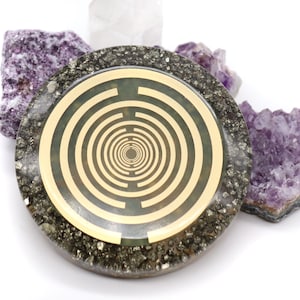 May include: A round, gold-coloured, geometric pattern is embedded in a circular, dark grey, stone-like disc. The disc is surrounded by small, purple crystals.
