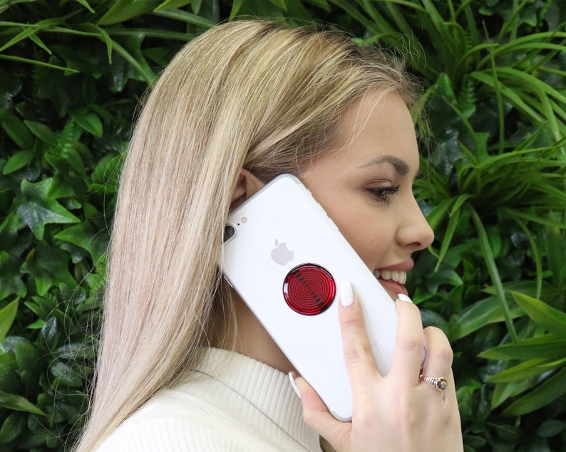 May include: A white smartphone featuring a circular accessory on the back with a red and black concentric circle pattern. The device has a silver Apple logo centred on the rear casing, positioned above the circular red and black graphic design.