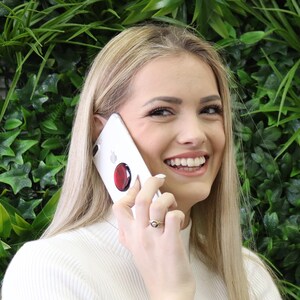 May include: A white smartphone featuring a circular red collapsible grip attached to the back. The device is held against the side of a face, showcasing the sleek white casing and the contrasting vibrant red accessory on the rear surface.