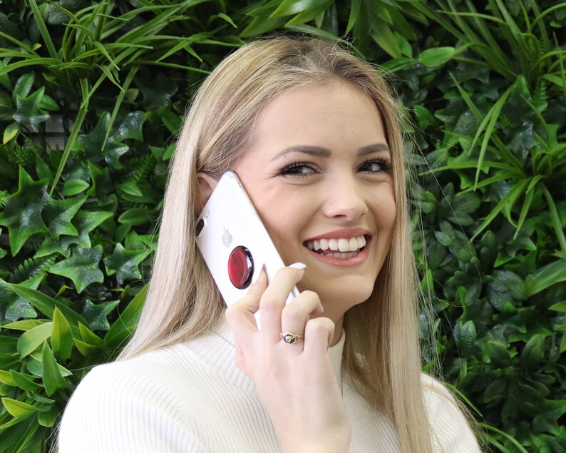 May include: A white smartphone featuring a circular red collapsible grip attached to the back. The device is held against the side of a face, showcasing the sleek white casing and the contrasting vibrant red accessory on the rear surface.