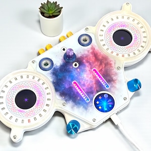 以下が含まれることがあります： 白を基調とした電子機器で、銀河をテーマにしたデザインです。複数のノブ、ボタン、円形のディスプレイがあり、カラフルなLEDライトが付いています。このデバイスにはUSBケーブルが付属しており、オーディオまたはビジュアルエフェクトに使用される可能性があります。
