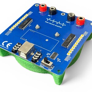 May include: A blue dual-channel amplifier circuit board with a green base. It features various electronic components, including a USB port, audio jacks, and terminal connectors. The board has text that reads "Dual channel amplifier".