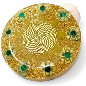Può includere: Un dispositivo di energia orgonica rotondo, giallo dorato, con un motivo geometrico centrale e dettagli a spirale. Incorpora pietre di malachite verde e glitter dorati, appoggiato su una pietra chiara.