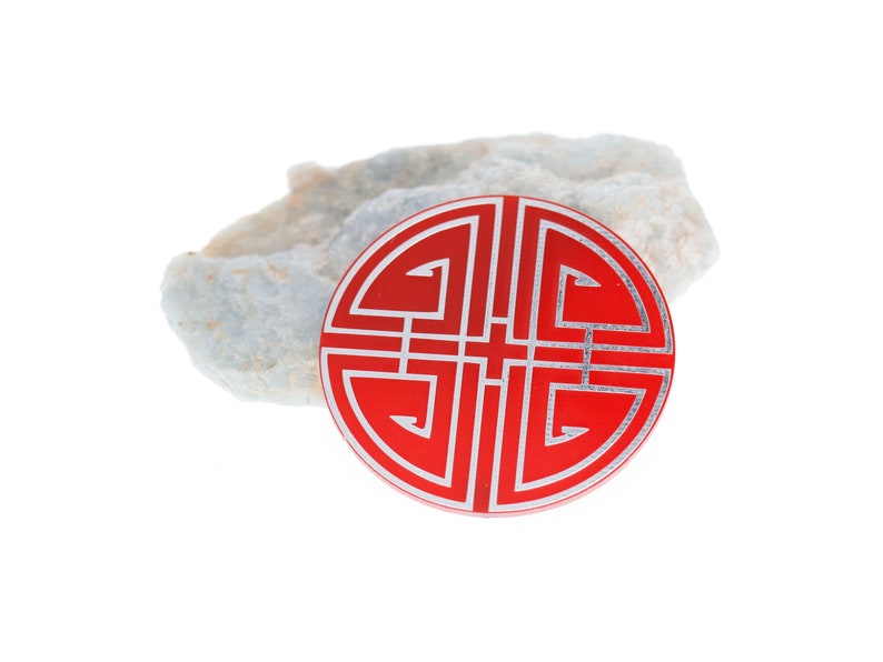 May include: A red circular charm with a silver design of a traditional Chinese knot. The charm is sitting on a piece of white rock.