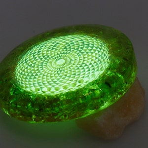 May include: A glowing green, oval-shaped object with a detailed geometric pattern in the center. The object is resting on a light-colored, textured surface. The edges of the object have a rough, crystalline appearance.