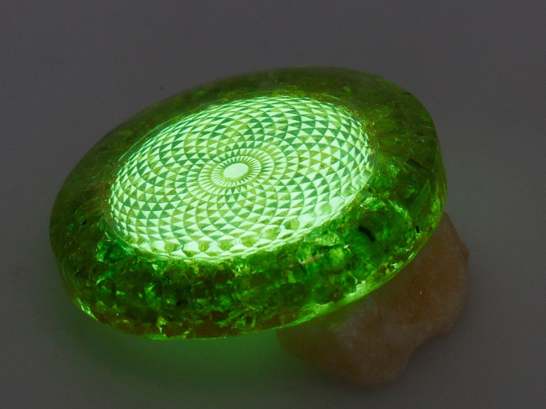 May include: A glowing green, oval-shaped object with a detailed geometric pattern in the center. The object is resting on a light-colored, textured surface. The edges of the object have a rough, crystalline appearance.