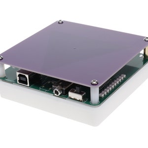 Op de afbeelding: Een close-up van een klein, vierkant, wit elektronisch apparaat met een paarse bovenkant. Het apparaat heeft een USB-poort, een kleine zwarte schakelaar en een groene printplaat die zichtbaar is onder de bovenkant.