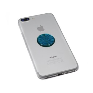 May include: A silver smartphone with a dual-lens camera system and an Apple logo on the back. A circular, metallic blue adhesive accessory with a concentric ring pattern is attached to the centre of the phone's rear casing.
