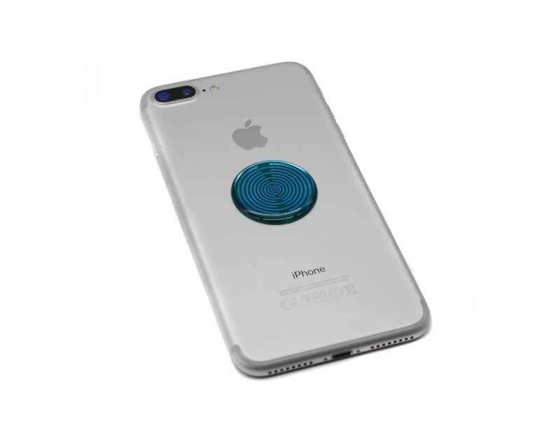 May include: A silver smartphone with a dual-lens camera system and an Apple logo on the back. A circular, metallic blue adhesive accessory with a concentric ring pattern is attached to the centre of the phone's rear casing.