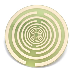 May include: A round, light green coaster with a concentric maze design in a lighter beige color. The maze pattern spirals inward from the outer edge to the center of the coaster. The coaster is set against a white background.