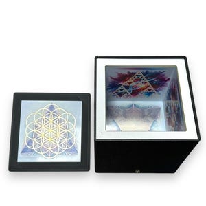 Op de afbeelding: Twee geometrische kunstwerken. Eén is een zwart omkaderde vierkant met een bloem des levens ontwerp. De andere is een zwarte doos met een geometrisch ontwerp binnenin. De doos heeft een witte rand en een doorzichtige bovenkant.