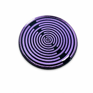 May include: A circular pin featuring a metallic purple finish with a black concentric maze pattern. The design consists of thin, repeating circular lines that radiate from a central point, creating a labyrinthine effect across the surface.