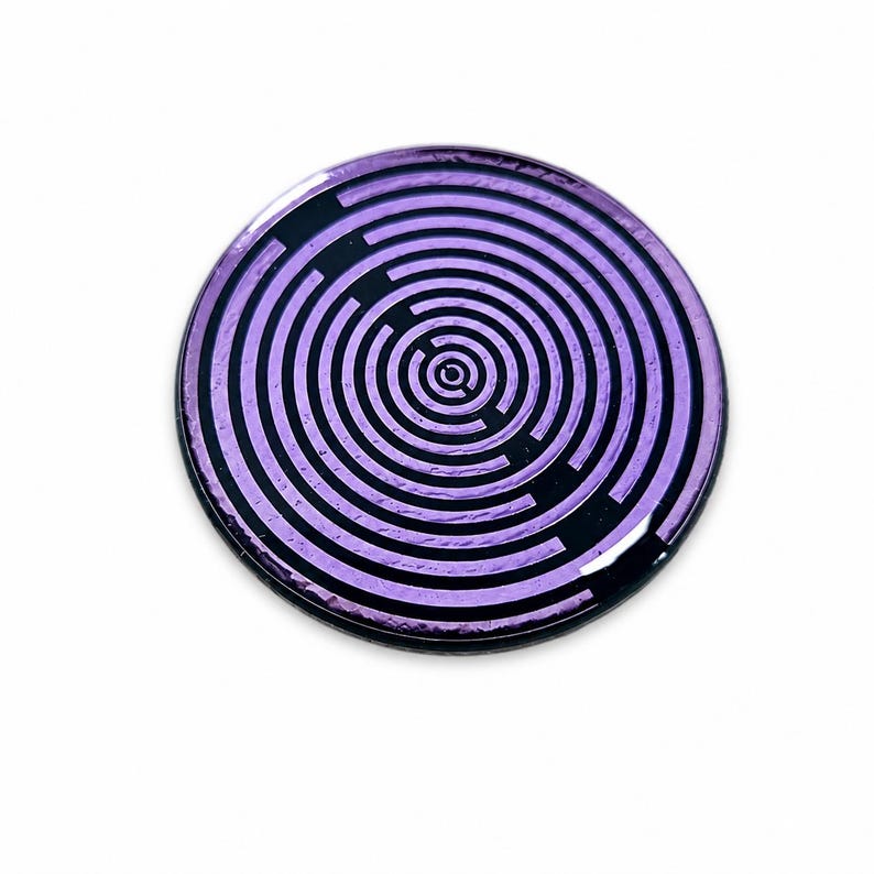 May include: A circular pin featuring a metallic purple finish with a black concentric maze pattern. The design consists of thin, repeating circular lines that radiate from a central point, creating a labyrinthine effect across the surface.