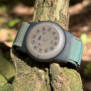 Puede incluir: Un reloj digital circular con una caja gris oscuro mate y una esfera translúcida que revela componentes electrónicos internos dispuestos en círculo. El reloj cuenta con una correa de nailon verde oliva texturizada con cierre de velcro.