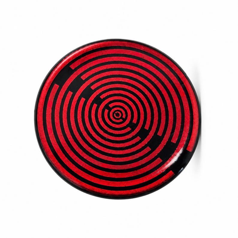 May include: A circular plate featuring a bold, high-contrast pattern of concentric red and black rings. The design consists of alternating thin red and black lines radiating from a central point, creating a geometric maze-like effect.