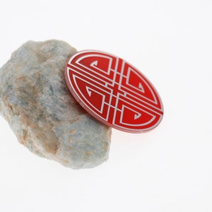 May include: A red circular charm with a white intricate design, resembling a knot, resting on a light gray rock.