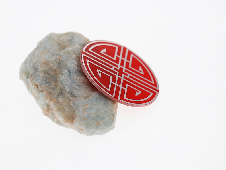 May include: A red circular charm with a white intricate design, resembling a knot, resting on a light gray rock.