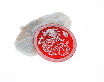 May include: A red circular disc with a white dragon design and a decorative border. The dragon is curled up with its head facing the left. The disc is sitting on a gray rock.