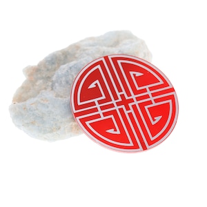 May include: A red circular charm with a silver design of a traditional Chinese knot. The charm is sitting on a piece of white rock.