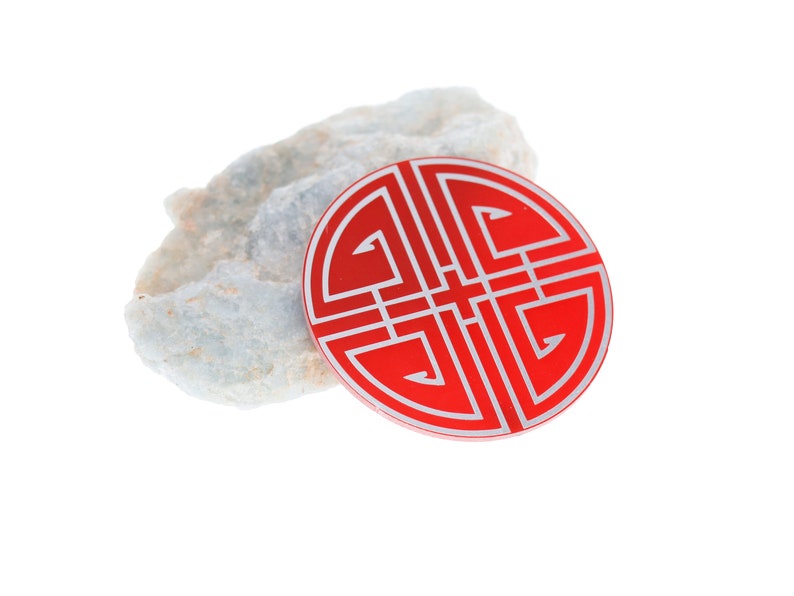 May include: A red circular charm with a silver design of a traditional Chinese knot. The charm is sitting on a piece of white rock.