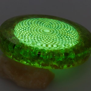 May include: A glowing green, circular object with a detailed geometric pattern on top. The object is made of a translucent material, with a lighter green center and a darker green outer ring. It rests on a light-colored, textured surface.