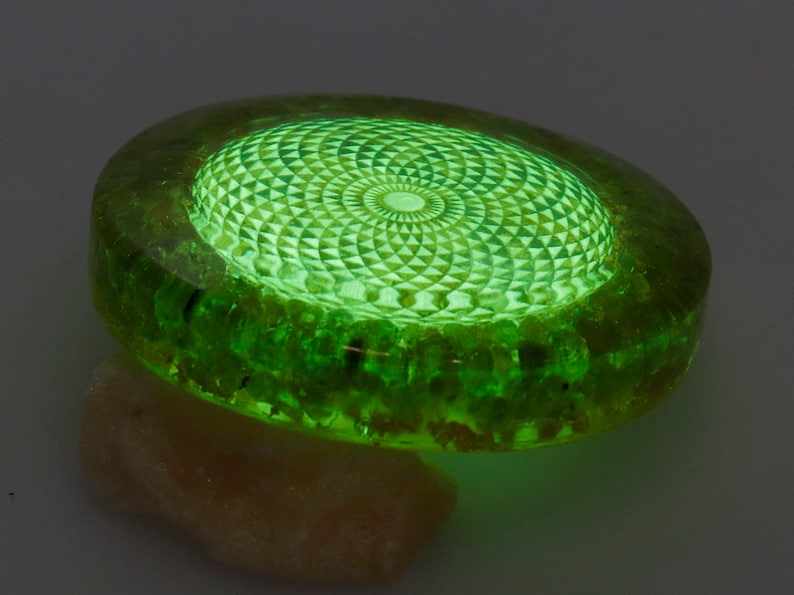 May include: A glowing green, circular object with a detailed geometric pattern on top. The object is made of a translucent material, with a lighter green center and a darker green outer ring. It rests on a light-colored, textured surface.